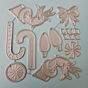 May include: A die-cut metal sheet featuring various Christmas-themed shapes. The shapes include a candy cane, ornaments, a bow, a snowflake, and other festive designs. The die-cut sheet is silver-colored and sits on a light blue surface.