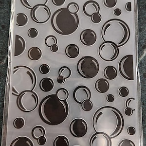 May include: A rectangular plastic embossing folder featuring a repeating pattern of various sized circles and bubbles. The design consists of raised black circular outlines and solid black circular shapes set against a translucent, clear background.