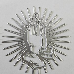 Op de afbeelding: Een zilverkleurige metalen stansvorm in de vorm van biddende handen, omringd door stralen. De handen zijn gedetailleerd met individuele vingers en een getextureerd oppervlak, wat een religieus of spiritueel thema cre&euml;ert.