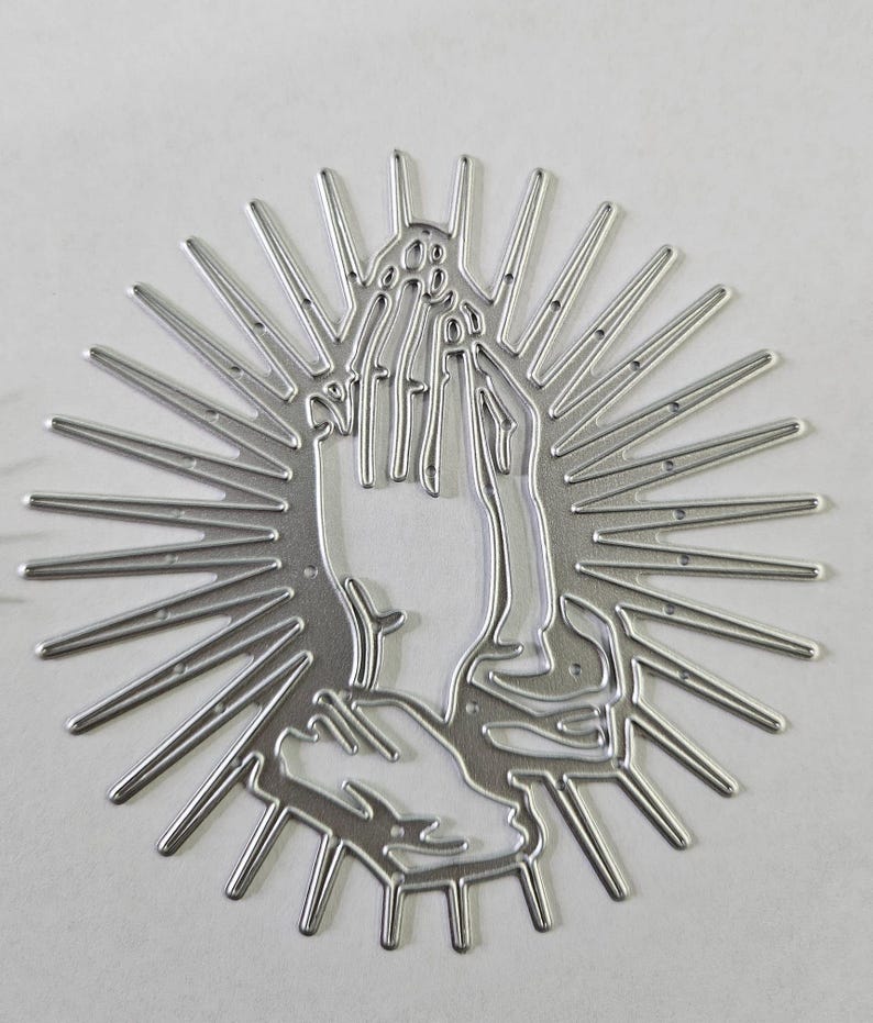 Op de afbeelding: Een zilverkleurige metalen stansvorm in de vorm van biddende handen, omringd door stralen. De handen zijn gedetailleerd met individuele vingers en een getextureerd oppervlak, wat een religieus of spiritueel thema cre&euml;ert.