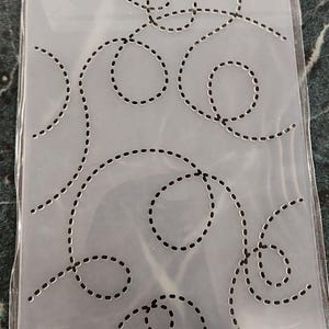 May include: A rectangular, semi-transparent plastic embossing folder featuring a repeating pattern of black dashed lines that form swirling, curved, and looping shapes across the entire surface of the clear, flexible crafting tool.