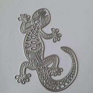 May include: A silver-colored metal die-cut in the shape of a lizard. The lizard features intricate cut-out patterns along its back, head, and tail, with small embossed dots lining the edges of its body, legs, and feet, creating a detailed silhouette.