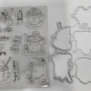 May include: Clear plastic sheet with stamps and dies for crafting. The stamps feature detailed line drawings of mugs filled with whipped cream, snowmen, and Christmas trees. Text includes "Mugs and kisses" and "Sugar 3 spice".