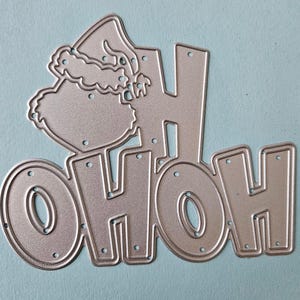 May include: A silver-colored metal die cut in the shape of the words "OHOH" with a Santa hat. The letters are blocky with small holes for attaching to a project. The hat is detailed with a fluffy brim.