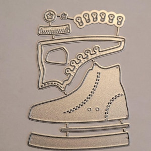 May include: A silver-colored metal die cut in the shape of an ice skate. The die cut includes details such as laces, eyelets, and a blade. Additional decorative elements include stars, circles, and a ruler.