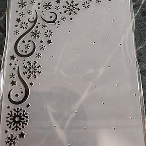 May include: A rectangular clear plastic embossing folder featuring a raised design of snowflakes, stars, and swirling lines concentrated along the left and top edges, with scattered small dots across the remaining surface area.