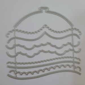 May include: A silver metal die cut with various decorative border designs. The die cut features a hanger-like top and several wavy and scalloped edges, perfect for paper crafting and scrapbooking projects.