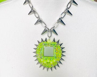 May include: A silver-toned chain necklace with large, pointed spikes. The pendant is a neon green heart shape with a square mirror in the center, surrounded by more spikes and small, clear embellishments. The necklace is displayed on a white surface.