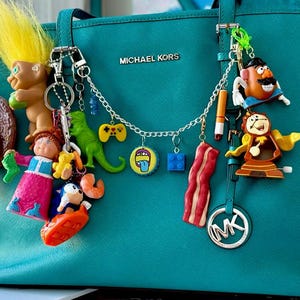 Op de afbeelding: Teal Michael Kors-draagtas versierd met een verscheidenheid aan kleurrijke sleutelhangers. Sleutelhangers zijn onder meer een hamburger, een trollenpop, een dinosaurus, een gamecontroller, een Lego-steen, een plakje spek en andere novelty-bedels.