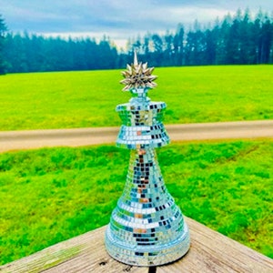 Könnte beinhalten: Eine silberne, verspiegelte Schachfigur in Form eines Königs mit einer sternförmigen Spitze. Die Figur steht auf einer Holzoberfläche mit einem grünen Grasfeld und Bäumen im Hintergrund.
