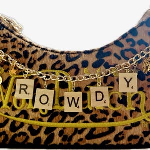 May include: A leopard print handbag with gold chain accents and charms. The word "ROWDY" is spelled out with wooden letter tiles. Additional charms include a cowboy hat, boot, and horseshoe. A yellow tassel hangs from the side.