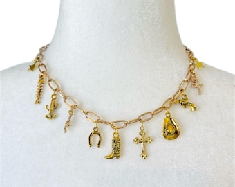 May include: Gold-toned charm necklace featuring a variety of Western-themed charms. The charms include a cowboy boot, horseshoe, cross, cactus, and a cowboy hat. The necklace is displayed on a white mannequin.