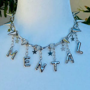 May include: Silver-toned choker necklace with spiked accents and dangling star charms. The word "MENTAL" is spelled out in individual silver letters hanging from the chain. The necklace is displayed on a white mannequin.