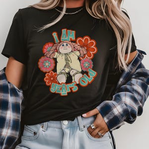 May include: Black t-shirt featuring a vintage doll graphic with the text "I AM YEARS OLD." The doll is surrounded by retro-style flowers in shades of pink, orange, and teal. The doll is wearing a yellow outfit.
