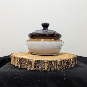 May include: A small brown and white ceramic pot with a lid. The pot has a rounded shape and two small handles on the sides. The pot is sitting on a wooden slice.