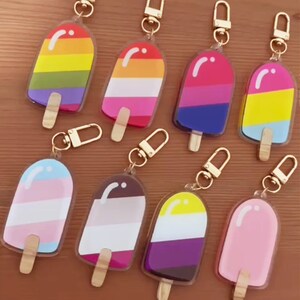 May include: Eight colorful popsicle keychains with gold metal rings. Each keychain features a different color combination of stripes, including rainbow, pink, blue, yellow, and purple.