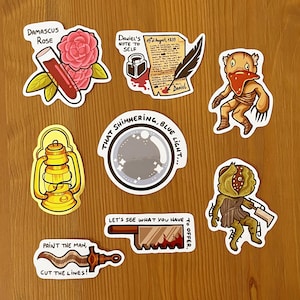 Może przedstawiać: Zestaw dziesięciu naklejek z różnymi ilustracjami, w tym różą, latarnią, kryształową kulą, piłą i stworzeniem z czerwonym szalikiem. Naklejki mają na sobie tekst, w tym "Damascus Rose", "Daniel's Note to Self", "That Shimmering, Blue Light...", "Paint the Man, Cut the Lines!" i "Let's See What You Have to Offer."