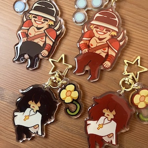 May include: Four colourful keychains featuring cartoon characters. Each keychain has a gold star charm and a flower charm. The characters are wearing different outfits and have different hairstyles.