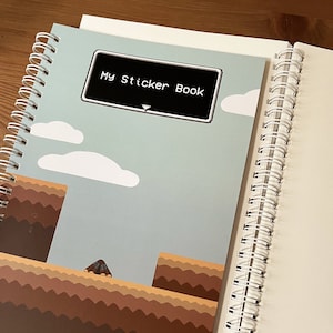 May include: A blue and brown spiral bound notebook with a white sticker on the front that says "My Sticker Book".