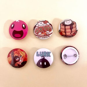 Pode incluir: Uma coleção de seis botões redondos e coloridos. Os botões apresentam vários designs, incluindo uma mancha rosa sorridente, um gato com comida, uma pilha de panquecas, um personagem com cabelo ruivo, a palavra "LURK" e uma parte traseira do botão.