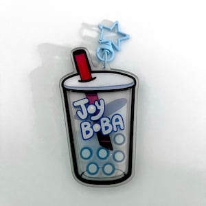 May include: A clear acrylic keychain in the shape of a boba tea cup. The cup features a white rim, a red straw, and the words "Joy Boba" in blue. It is attached to a blue star-shaped clasp.
