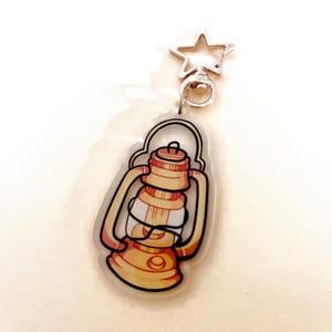 May include: A clear acrylic keychain featuring a cartoon illustration of a vintage lantern in shades of yellow, orange, and white, outlined in black. The keychain has a silver star-shaped clasp.