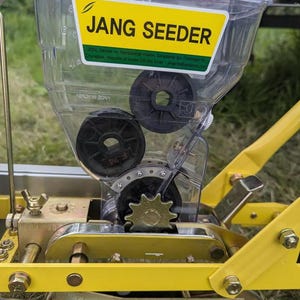 Könnte beinhalten: Nahaufnahme einer gelben und silbernen Jang-Sämaschine. Die Sämaschine hat einen transparenten Kunststoff-Saatbehälter mit schwarzen Zahnrädern und einem gelben Etikett mit der Aufschrift "JANG SEEDER". Die Sämaschine ist zum Pflanzen von Samen konzipiert.