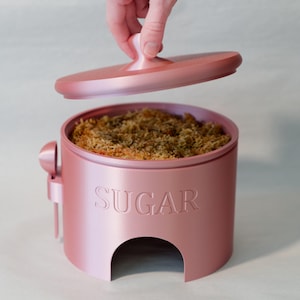 May include: A pink ceramic sugar dispenser with a lid and a spoon. The dispenser has the word "SUGAR" embossed on the front.
