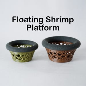 May include: Two floating shrimp platforms. One is olive green, the other is brown. Both have a porous, honeycomb-like design and a dark gray rim. The text "Floating Shrimp Platform" is above the platforms.