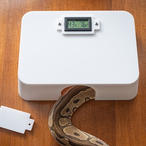 May include: A white plastic reptile enclosure with a digital temperature and humidity gauge displaying 43% and 76 degrees Fahrenheit. A brown and tan snake is partially visible entering the enclosure.