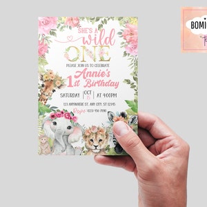 May include: A pink and gold first birthday invitation with a jungle animal theme. The invitation features a giraffe, elephant, lion, and zebra with floral accents. The text reads "She's a wild one Annie's 1st Birthday Saturday Oct 15 at 4:00pm 123 Anywhere St. Any City, ST 12345 Repu- (023) 456-7890"