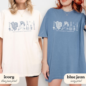May include: Two t-shirts, one ivory and one blue jean, featuring a white graphic design. The design includes illustrations of a bouquet, champagne bottle, and other celebratory elements. Text on the shirts reads "EMILY'S BACHELORETTE + CHARLESTON 2015".