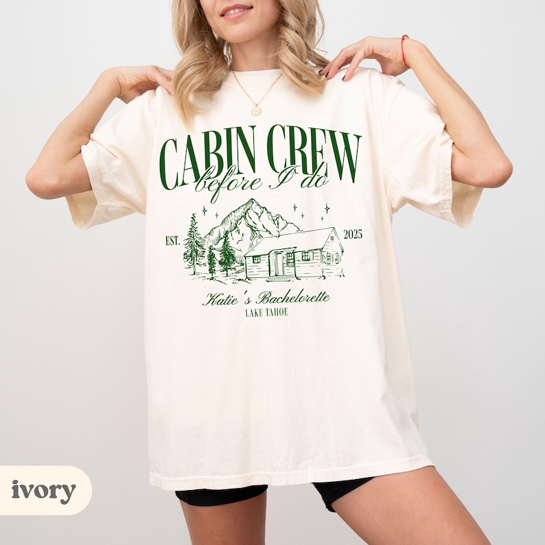 May include: Ivory t-shirt featuring the text "CABIN CREW before I do" in dark green. A cabin, mountains, and trees graphic is below the text. The text "Katie's Bachelorette LAKE TAHOE" is also included on the shirt.
