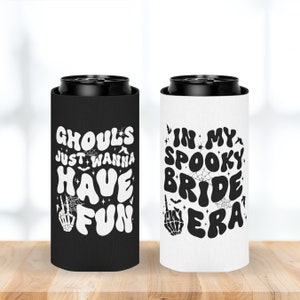 May include: Two black and white can coolers with Halloween themed designs. The cooler on the left says "ghouls just wanna have fun" with a skeleton hand and bats. The cooler on the right says "in my spooky bride era" with a skeleton hand and bats.