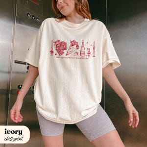 May include: Ivory-colored t-shirt with a chili-red graphic design. The design includes illustrations of a bouquet, champagne bottle, and other celebratory items. Text on the shirt reads "EMILY'S BACHELORETTE CHARLESTON 2023".