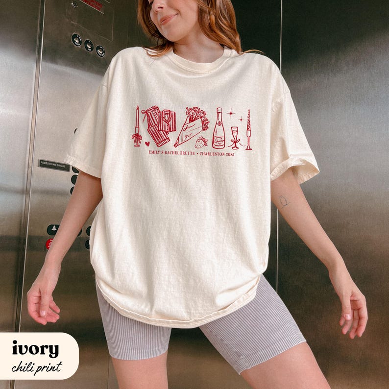 May include: Ivory-colored t-shirt with a chili-red graphic design. The design includes illustrations of a bouquet, champagne bottle, and other celebratory items. Text on the shirt reads "EMILY'S BACHELORETTE CHARLESTON 2023".