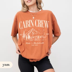 May include: A rust-coloured t-shirt featuring the text "CABIN CREW" in white, above a cabin and mountain graphic. The shirt also displays "Katie's Bachelorette LAKE TAHOE" in white text, ideal for a bachelorette party.