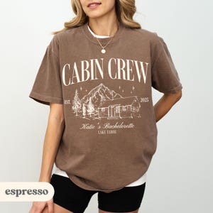 May include: Espresso-coloured t-shirt featuring the words "CABIN CREW" above a cabin and mountain graphic. Below the graphic is "Katie's Bachelorette LAKE TAHOE" and the year "2025". The shirt has short sleeves and a crew neck.