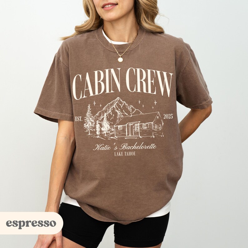 May include: Espresso-coloured t-shirt featuring the words "CABIN CREW" above a cabin and mountain graphic. Below the graphic is "Katie's Bachelorette LAKE TAHOE" and the year "2025". The shirt has short sleeves and a crew neck.