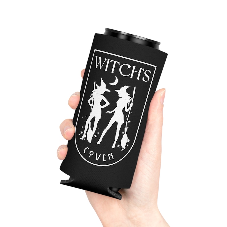 Puede incluir: Un enfriador de latas negro con el texto "WITCH'S COVEN" en blanco. El dise&ntilde;o presenta dos siluetas de brujas, una luna creciente y estrellas. El enfriador es sostenido por una mano.