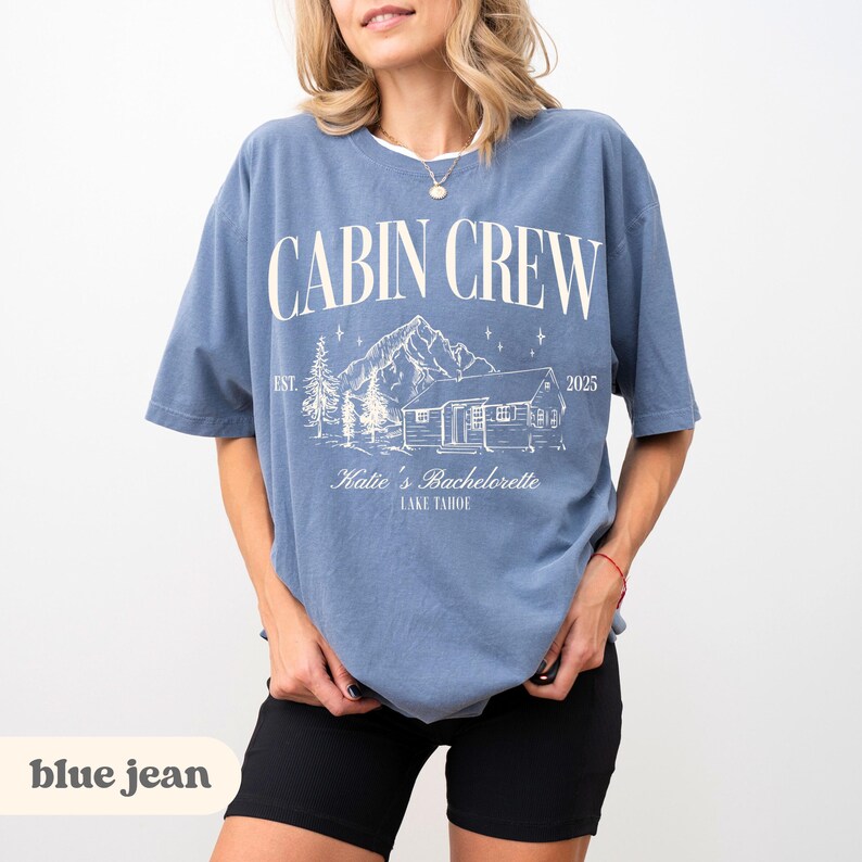 May include: A blue t-shirt featuring the words "CABIN CREW" in white. The shirt has a graphic of a cabin, mountains, and trees. The text "Katie's Bachelorette LAKE TAHOE" is printed below the graphic.