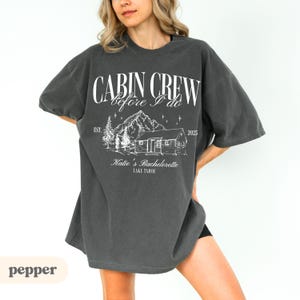 May include: Dark grey t-shirt featuring the text "CABIN CREW before I do" above a cabin and mountain graphic. The text "Katie's Bachelorette LAKE TAHOE" is below the graphic. The year "2025" is also printed on the shirt.