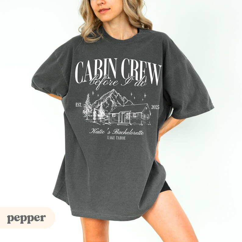 May include: Dark grey t-shirt featuring the text "CABIN CREW before I do" above a cabin and mountain graphic. The text "Katie's Bachelorette LAKE TAHOE" is below the graphic. The year "2025" is also printed on the shirt.