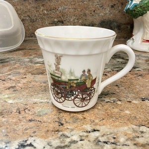 Può includere: Tazza in porcellana bianca con bordo dorato. La tazza presenta un'illustrazione vintage di una carrozza trainata da cavalli con due persone all'interno. Il testo "Daimler 1886" è stampato sul fondo della tazza.