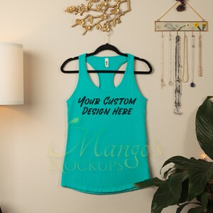 Op de afbeelding: Een turquoise tanktop met de tekst "YOUR CUSTOM DESIGN HERE" in zwart schrift. De tanktop hangt aan een zwarte hanger tegen een neutrale muur. De tanktop is een mock-up voor aangepaste ontwerpen.