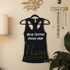 Op de afbeelding: Een zwart tanktop met een ronde hals en de tekst "Your Custom Design Here" gedrukt op de voorkant. Het tanktop hangt aan een hanger tegen een lichtbeige muur.