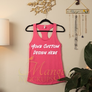 Op de afbeelding: Roze tanktop zonder mouwen met een ronde hals en racerback. Het tanktop hangt aan een hanger tegen een witte muur. De tekst "Your Custom Design Here" is gedrukt op de voorkant van het tanktop.