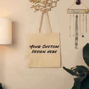May include: A beige canvas tote bag with black text that reads "Your Custom Design Here". The bag is hanging on a wall with a gold decorative branch above it.