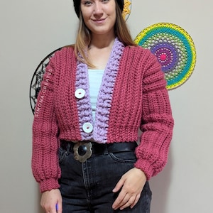 Pu&ograve; includere: Un cardigan di maglia rosa e viola con chiusura a bottone. Il cardigan ha una trama a coste e una lunghezza corta. La modella indossa il cardigan con una cintura nera e jeans neri.