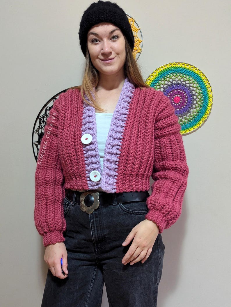 Pu&ograve; includere: Un cardigan di maglia rosa e viola con chiusura a bottone. Il cardigan ha una trama a coste e una lunghezza corta. La modella indossa il cardigan con una cintura nera e jeans neri.